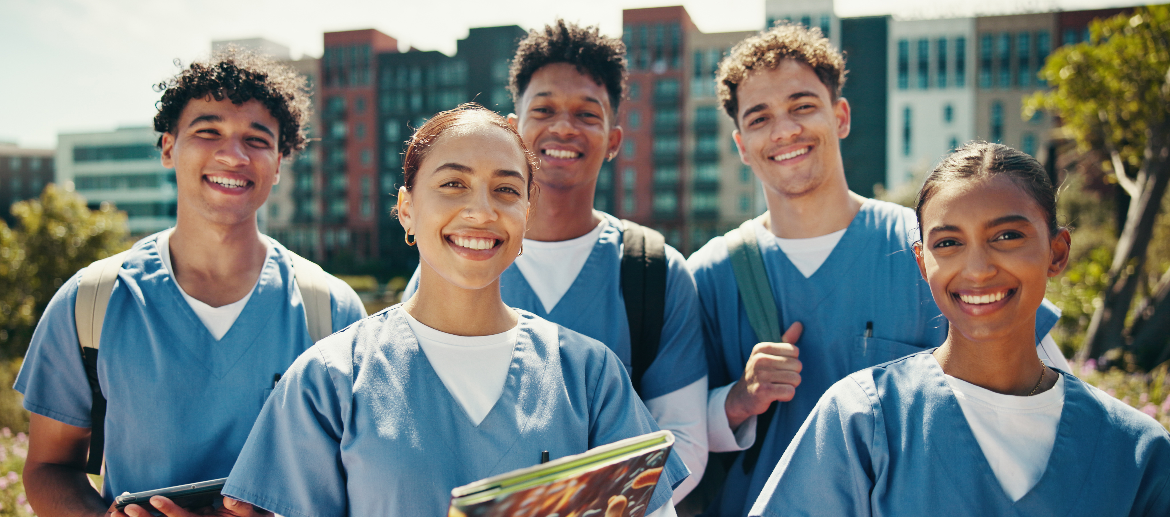 Group of diverse Licensed Vocational Nursing (LVN) students in blue scrubs smiling outdoors on campus with city buildings in the background